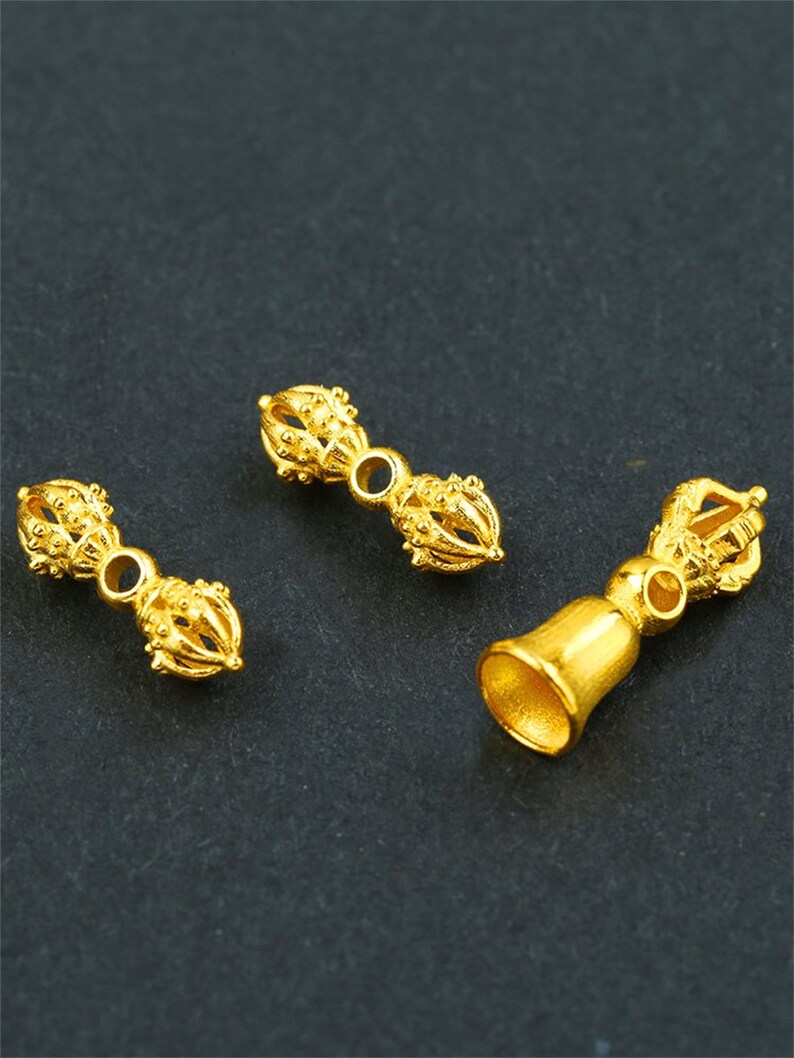 May include: Three gold-colored metal charms in the shape of a bell and two double-sided prayer wheels. The prayer wheels have intricate designs and are connected by a small ring.