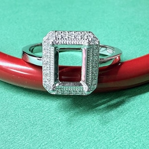 Puede incluir: Un anillo de plata con un engaste cuadrado rodeado de pequeños diamantes. El anillo está sobre una superficie roja y verde.