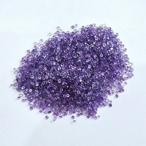 May include: A large pile of small, round, purple amethyst gemstones. The gemstones are scattered on a white surface.