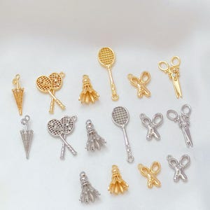 May include: A collection of gold and silver-toned charms in various shapes. The charms include badminton rackets, shuttlecocks, scissors, and umbrellas. The charms are arranged on a white surface, showcasing their intricate details and designs.