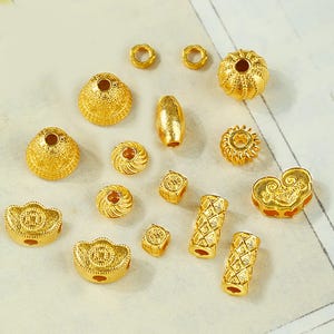 May include: An assortment of gold-coloured beads in diverse forms, such as round, cylindrical, and heart-shaped. The beads feature detailed patterns and textures, ideal for jewellery making or crafts. The selection includes small, medium, and large sizes.