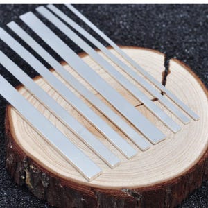 May include: A collection of six flat, rectangular silver-coloured metal strips of varying widths arranged in a parallel diagonal formation. Each strip features a smooth, reflective surface and straight edges, showcasing a uniform metallic finish.