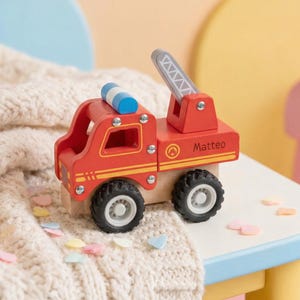 Può includere: Un camion dei pompieri giocattolo in legno rosso con una scala argentata, ruote nere e il nome "Matteo" stampato sul lato. Il camion ha una barra luminosa blu e bianca sulla parte superiore. Il giocattolo è su un tavolo bianco con bordi blu.