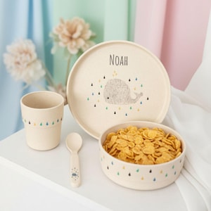 Pode incluir: Um conjunto de louça infantil bege com um design de baleia. O conjunto inclui um prato com o nome "NOAH", uma tigela cheia de cereais, um copo e uma colher. O prato e a tigela têm uma ilustração de baleia e detalhes coloridos de gotas.