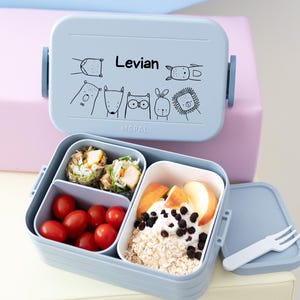 Op de afbeelding: Een blauwe lunchbox met de gepersonaliseerde naam "Levian" en een ontwerp van cartoondieren. De lunchbox heeft drie compartimenten en is gevuld met eten, waaronder fruit, groenten en een wrap.