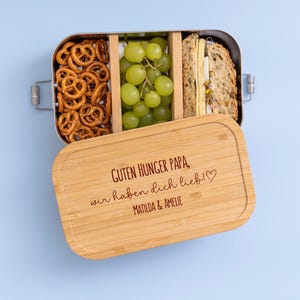 May include: A stainless steel lunchbox with a bamboo lid, filled with pretzels, grapes, and a sandwich. The lid is engraved with the text "GUTEN HUNGER PAPA, wir haben dich lieb!" and the names "MATILDA & AMELIE".