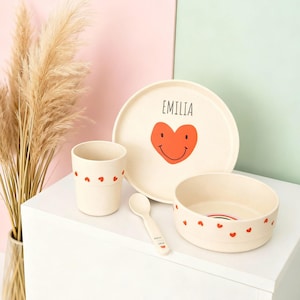 Pode incluir: Um conjunto de jantar infantil bege com prato, tigela, xícara e colher. O prato apresenta um coração vermelho com um rosto sorridente e o nome "EMILIA". A xícara e a tigela têm detalhes de coração vermelho. A colher tem o nome "EMILIA".