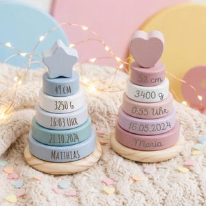 Può includere: Due giocattoli impilabili in legno dai colori pastello. Uno ha una stella, l'altro un cuore. Ognuno ha dettagli incisi: altezza in cm, peso in grammi, ora, data e un nome. I giocattoli poggiano su una base di legno.