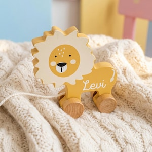 Op de afbeelding: Een gele houten trekfiguur in de vorm van een leeuw met een witte manen en gezicht. De leeuw heeft een lachend gezicht en de naam "Levi" staat op de zijkant. Het speelgoed ligt op een crèmekleurige gebreide deken.