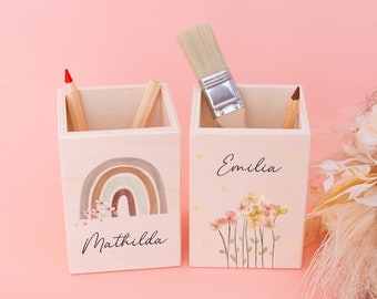 Op de afbeelding: Twee houten potloodhouders met gepersonaliseerde namen, "Matilda" en "Emilia". De ene houder heeft een regenboogontwerp met bloemen, terwijl de andere een bloemenontwerp met sterren heeft. Beide houders staan op een roze oppervlak.