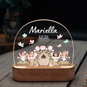 Peut inclure: Une veilleuse en bois avec un dôme en acrylique transparent représentant une scène féerique avec quatre fées, des fleurs et des oiseaux. Le dôme est éclairé par le bas, créant une lueur chaleureuse. Le nom "Mariella" et la date "16.03.2024" sont inscrits sur le dôme.