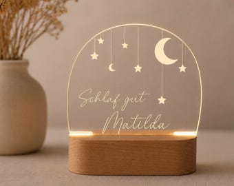 May include: An arched acrylic night light with a wooden base, featuring an engraved crescent moon, stars, and the text "Schlaf gut Matilda" illuminated in white. The design includes hanging celestial elements suspended from the top arch.
