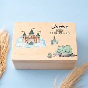 Puede incluir: Caja de recuerdos de madera con un acabado de madera clara. La tapa presenta una ilustración de acuarela de un castillo en las nubes, un dragón verde y un pequeño cactus. La caja está etiquetada con "Justus 10.03.2024 01:11 Uhr - 3810 g - 51 cm".