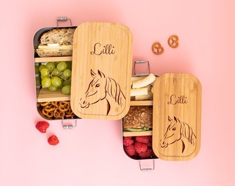 Op de afbeelding: Twee bruine houten lunchboxen met een paardenontwerp en de naam "Lilli" gegraveerd op de deksel. De lunchboxen zijn gevuld met eten, waaronder een sandwich, druiven, pretzels en frambozen.