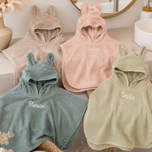 Könnte beinhalten: Vier Kapuzenponchos aus Frottee in Beige, Altrosa, Salbeigrün und Olivgrün. Jeder Poncho hat zwei kleine Ohren an der Kapuze und ist mit einem Namen bestickt: Elias, Malia, Florian oder Emilia in weißer Schreibschrift.