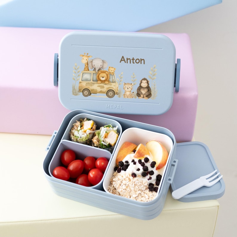 May include: A blue lunchbox with a safari animal design and the name "Anton" on the lid. The lunchbox is open and contains a variety of food, including fruit, vegetables, and a sandwich.