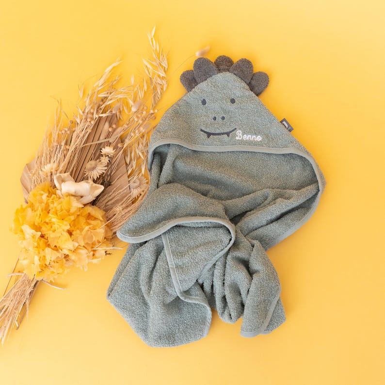 May include: A green hooded towel with a dinosaur head design. The towel has a gray border and the words "Benno" embroidered on the hood.