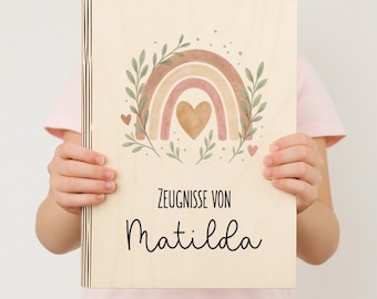 Pode incluir: Uma caixa de madeira com um design de arco-íris e coração. O texto "Zeugnisse von Matilda" está impresso na caixa.