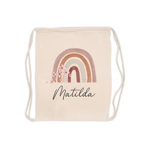 May include: A beige drawstring backpack featuring a watercolor rainbow design in shades of brown, tan, and pink, with delicate floral accents. The name "Matilda" is written in a cursive font below the rainbow.