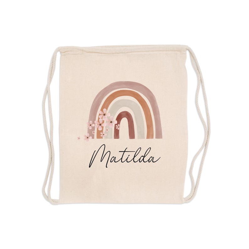 May include: A beige drawstring backpack featuring a watercolor rainbow design in shades of brown, tan, and pink, with delicate floral accents. The name "Matilda" is written in a cursive font below the rainbow.