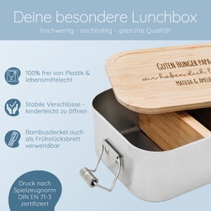 May include: A stainless steel lunchbox with a bamboo lid. The lid is engraved with text. The lunchbox has a metal handle and is labeled as food-safe and plastic-free. Text on the lid reads "GUTEN HUNGER PAPA wir haben dich l MATILDA & AMELIE".