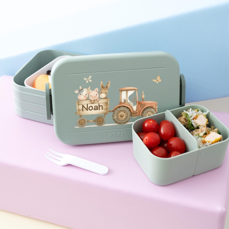 May include: A green and blue lunchbox with a tractor and farm animals on the front. The lunchbox is labeled "Noah".