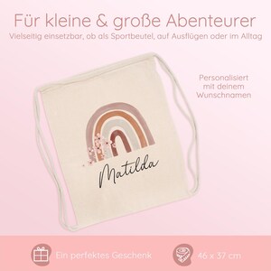 May include: A beige drawstring backpack with a watercolor rainbow design in shades of brown and pink, with the name "Matilda" in cursive. The bag measures 46 cm x 37 cm. Text on the bag says "F&uuml;r kleine & gro&szlig;e Abenteurer" and "Personalisiert mit deinem Wunschnamen."