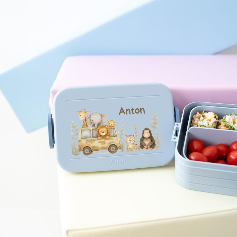May include: A blue lunchbox with a safari animal design and the name Anton on the front. The lunchbox is open and shows a compartment with a small sandwich and a compartment with cherry tomatoes.