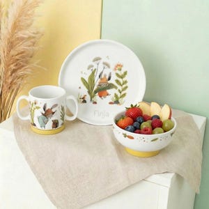 Könnte beinhalten: Ein weißes Keramik-Kindergeschirr-Set mit einem Hasen- und Blumendesign. Das Set enthält eine Tasse, einen Teller und eine Schüssel mit gelbem Boden. Auf dem Teller steht der Name "Finja". Die Schüssel ist mit frischem Obst gefüllt.