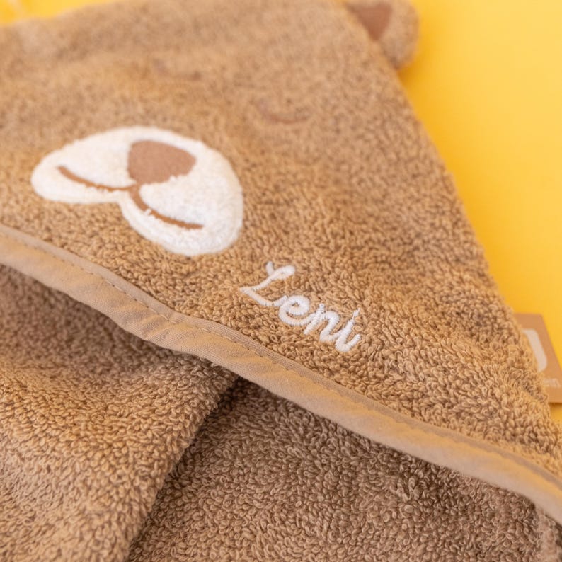 May include: A brown hooded towel with a white embroidered animal face and the name "Leni" stitched on the hood.