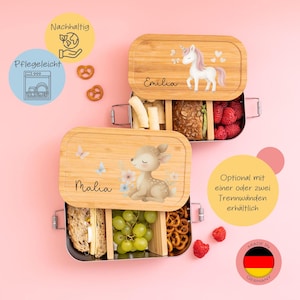 Può includere: Due contenitori per il pranzo in acciaio inossidabile con coperchi in bambù. I coperchi presentano un unicorno e un cervo con i nomi "Emilia" e "Malia" scritti su di essi. I contenitori per il pranzo sono pieni di cibo, tra cui frutta, pane e pretzel. Il testo "Optional mit einer oder zwei Trennwänden erhältlich" è scritto in tedesco sul lato destro dell'immagine. Il testo "MADE IN GERMANY" è scritto in nero su una bandiera rosso, giallo e nero.