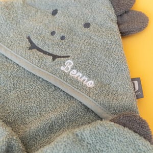 May include: A green hooded towel with a smiling face and the name "Benno" embroidered on it. The towel has gray ears and is made of soft, absorbent material.