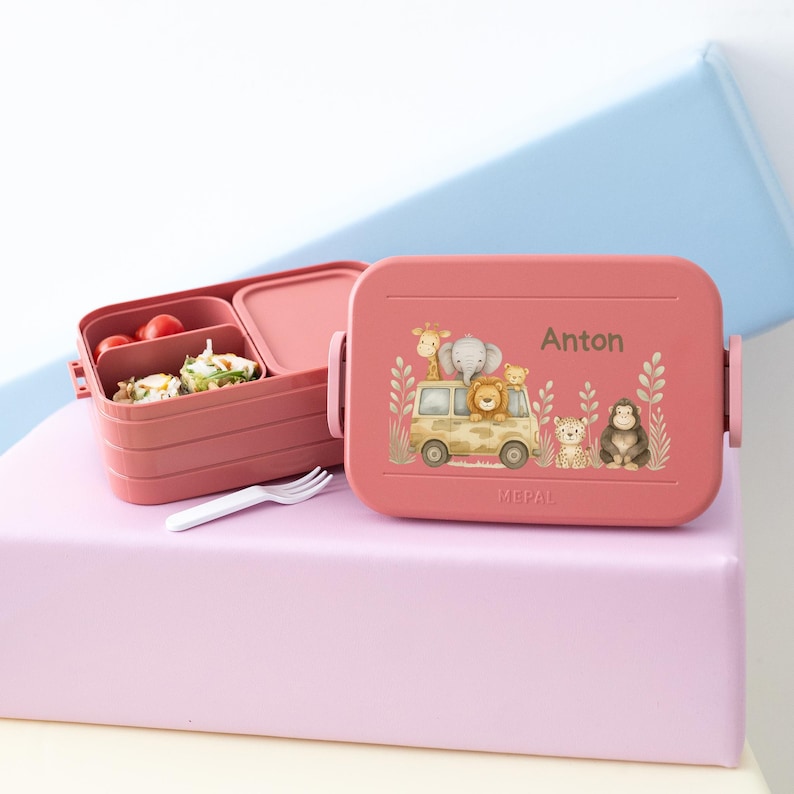 May include: Pink lunchbox with a safari animal design and the name Anton on the front. The lunchbox has three compartments and is made by Mepal.