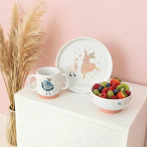 Peut inclure: Un ensemble de vaisselle pour enfants en céramique blanche comprenant une assiette, un bol et une tasse. L'assiette et la tasse présentent des illustrations d'une ballerine et d'un cygne. Le bol est rempli de fruits frais. Le nom "Mathilda" est imprimé sur l'assiette.