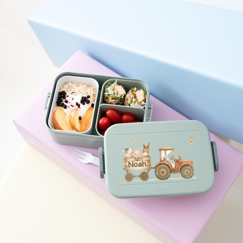 May include: A green lunchbox with a tractor design and the name "Noah" on the side. The lunchbox has three compartments and is filled with food, including fruit, yogurt, and a rice dish.