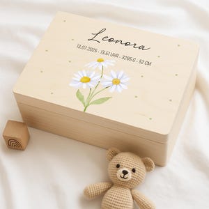 Può includere: Un cofanetto in legno chiaro con un design di margherite ad acquerello e il nome "Leonora" in corsivo. La scatola include anche la data, l'ora, il peso e l'altezza del bambino. Un piccolo blocchetto di legno e un orsacchiotto all'uncinetto sono accanto.