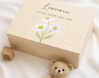 May include: A light-colored wooden keepsake box with a watercolor daisy design and the name "Leonora" in script. The box also includes the date, time, weight, and height of the baby. A small wooden block and a crocheted teddy bear are next to the box.