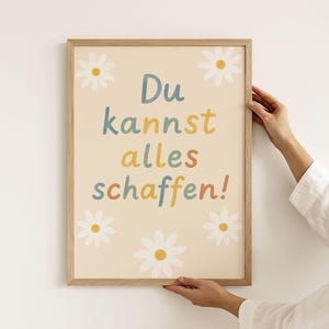 Könnte beinhalten: Ein gerahmter Druck mit einem hellbeigen Hintergrund und dem Text "Du kannst alles schaffen!" in bunten Buchstaben. Der Druck ist mit weißen Gänseblümchen verziert.