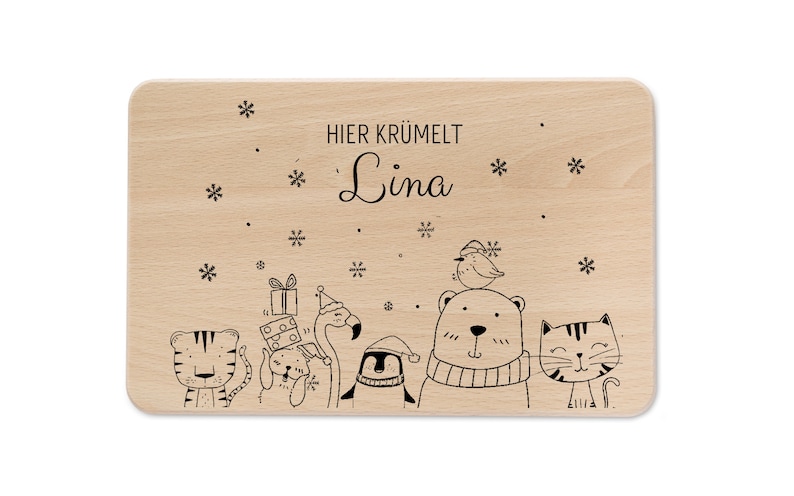 Pode incluir: Uma t&aacute;bua de cortar de madeira com uma ilustra&ccedil;&atilde;o em preto e branco de animais e flocos de neve. O texto "HIER KR&Uuml;MELT Lina" est&aacute; gravado na t&aacute;bua.