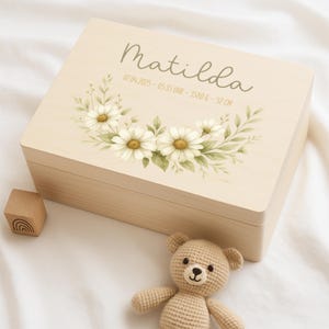 Può includere: Una scatola di legno per ricordi con una finitura in legno chiaro. Il coperchio presenta un motivo floreale con margherite bianche e foglie verdi. Il testo "Matilda" è stampato sul coperchio, insieme alla data "07.04.2023", l'ora "05:35 Uhr", il peso "3580 g" e la lunghezza "52 cm".