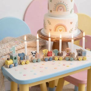 May include: A birthday cake with rainbow decorations sits on a wooden stand. A toy train with animal-themed cars holds lit candles. The train cars feature a number 1, an elephant, a panda, a bear, a lion, and a dinosaur. The table is white with a blue trim.