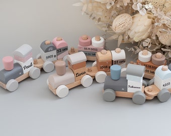 May include: A set of five wooden toy trains, each with a different name and birth information. The trains are painted in various shades of grey, pink, and brown. The trains are arranged in a line on a grey surface.