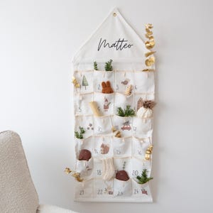 Peut inclure: Un calendrier de l'Avent en tissu crème avec 24 poches numérotées, chacune contenant de petites décorations. Le haut porte le nom "Matteo" en écriture cursive. Des arbres miniatures, des animaux et des brins de verdure ornent les poches. Une branche d'eucalyptus dorée est sur le côté.