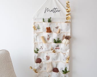 May include: A cream-colored fabric advent calendar with 24 numbered pockets, each containing small decorative items. The top features the name "Matteo" in script. Embellishments include miniature trees, animals, and sprigs of greenery. A gold-colored eucalyptus branch is on the side.