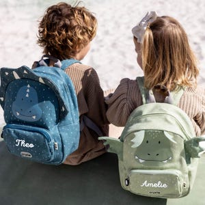 Könnte beinhalten: Zwei Kinder mit Rucksäcken im Dinosaurier-Design. Einer ist blau mit einem lächelnden Dinosauriergesicht und dem Namen "Theo". Der andere ist grün mit einem Drachengesicht und dem Namen "Amelie". Beide haben eine kleine Vordertasche.