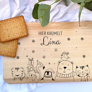 Pode incluir: Uma t&aacute;bua de cortar de madeira com o texto "HIER KR&Uuml;MELT Lina" e uma ilustra&ccedil;&atilde;o de inverno de animais, incluindo um pinguim, um flamingo, um urso, um gato e um tigre.