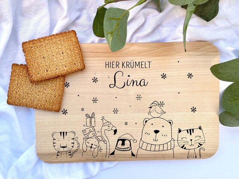 Pode incluir: Uma t&aacute;bua de cortar de madeira com o texto "HIER KR&Uuml;MELT Lina" e uma ilustra&ccedil;&atilde;o de inverno de animais, incluindo um pinguim, um flamingo, um urso, um gato e um tigre.