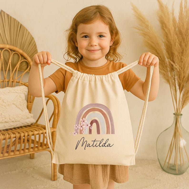 May include: A beige drawstring backpack featuring a watercolor rainbow design in shades of purple, brown, and blue, with floral accents. The name "Matilda" is written in a cursive font below the rainbow. The bag has cream-colored drawstrings.
