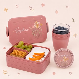 Pode incluir: Uma lancheira retangular rosa empoeirado com um recipiente redondo para lanches a condizer, ambos com o nome Sophia e uma ilustração floral. A lancheira aberta contém uvas verdes e bolachas, enquanto o recipiente contém mirtilos.
