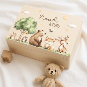 Op de afbeelding: Een houten bewaardoos met een lichtbruine afwerking. De deksel heeft een fantasierijke bos scène met een beer, vos, hert, eekhoorn en egel. De doos is gepersonaliseerd met de naam "Noah" en de datum "26.03.2025".